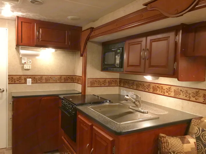 Step inside a clean RV rental kitchen, complete with dark wood cabinetry, stove, sink, and built-in microwave.. Forest River Wildwood 2011