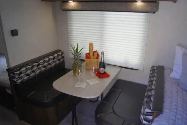RV rental interior: a dining nook with dark benches and a light table, set with fruit, bread, and drinks. A bed is nearby.. Heartland Other 2018