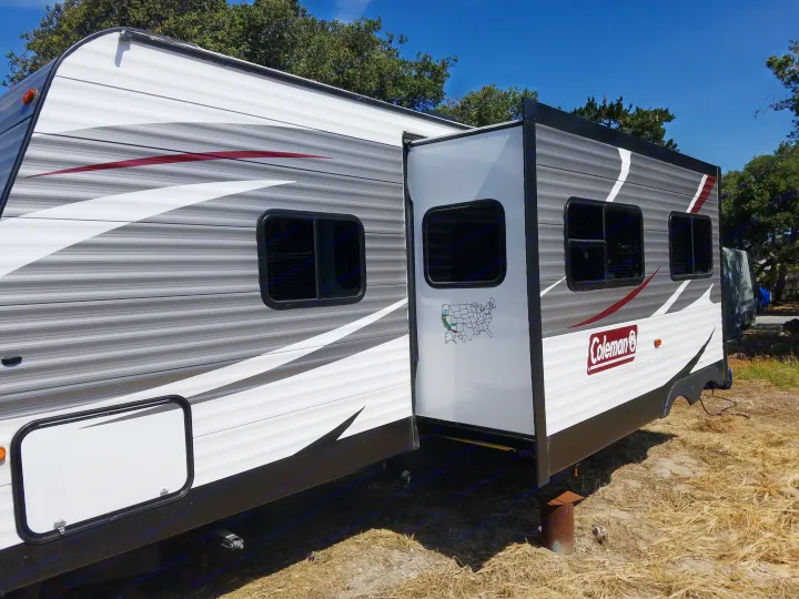A gray and white Coleman RV rental, with a slide-out extended, sits parked on dry ground under a blue sky.. Coleman 285bh 2017