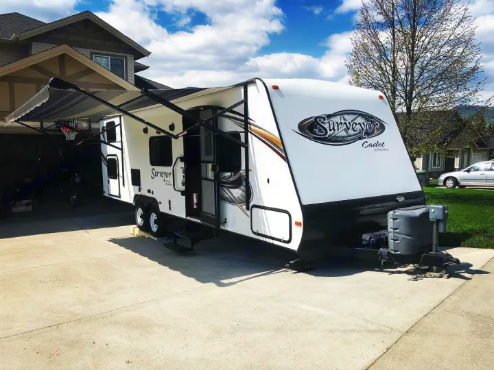 A white Surveyor Cadet travel trailer with its awning extended is parked on a concrete driveway. Perfect for your next RV rental.. Forest River Surveyor 2014