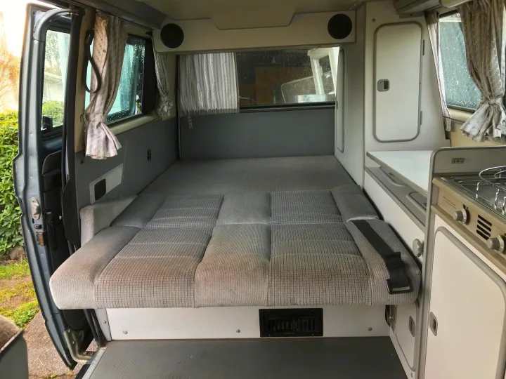 Inside this RV rental: a cozy grey patterned bed (cushions laid flat) and a compact kitchen. Ideal for road trips.. Volkswagen Westfalia 1989