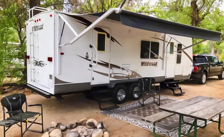 This RV rental travel trailer is set up at a leafy campsite, complete with an awning, picnic table, and chairs for outdoor relaxation.. Forest River Wildwood 2011