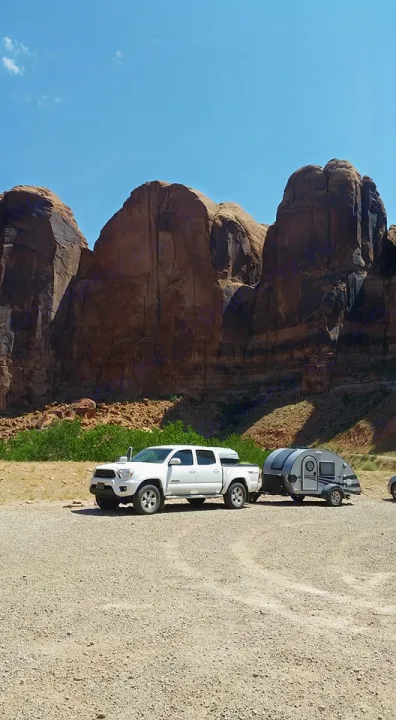 A white pickup truck tows a compact RV rental teardrop trailer on gravel, against towering desert cliffs under a blue sky.. Little Guy Silver Shadow 2016
