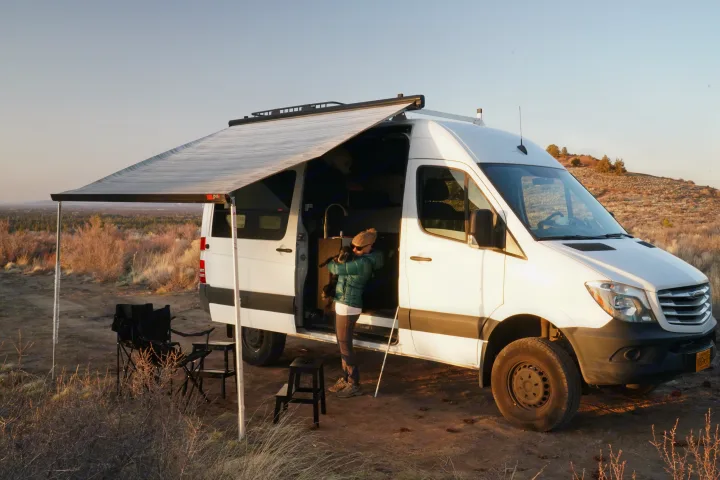 An RV rental camper van with an awning extended is parked in nature. A person holds a dog by the open side door.. Freightliner Sprinter 2016