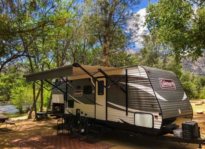 Riverside RV rental: A grey Coleman travel trailer with awning, set up in a shaded campsite by a flowing river.. Coleman Lantern 2018