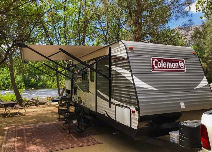 Riverside RV rental campsite features a travel trailer with an extended awning, outdoor rug, and picnic area by the water.. Coleman Lantern 2018