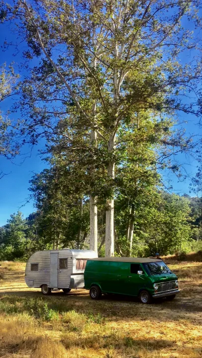 Green vintage van with silver camper, an ideal RV rental. Parked in a sunny field among tall trees.. Shasta Oasis 1959