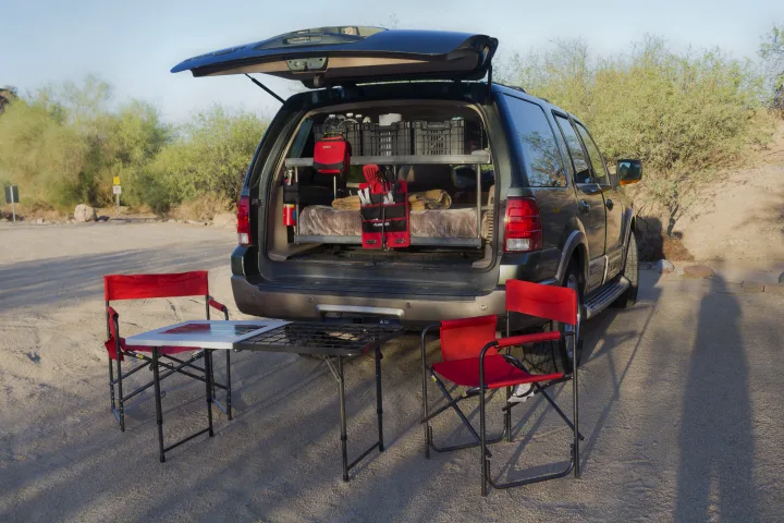SUV RV rental setup: Open trunk displays a bed, storage, and camping gear. Red chairs and a table are ready outdoors.. Ford Expedition Camper Van Roadtrip Camper Van 2003