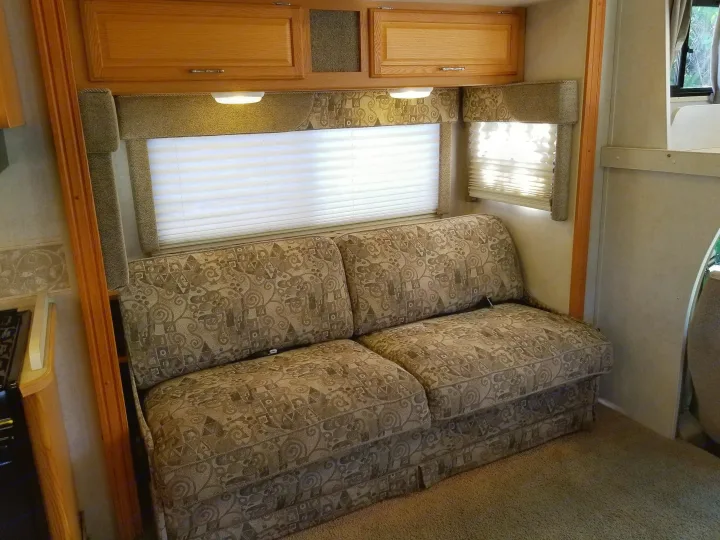 Cozy RV rental interior featuring a patterned sofa, window with blinds, and overhead storage cabinets, ideal for travelers.. Winnebago Outlook 2006