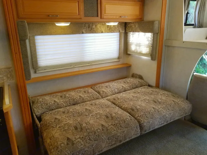 RV rental interior featuring a versatile sofa bed, wooden overhead cabinets, and bright windows with blinds.. Winnebago Outlook 2006