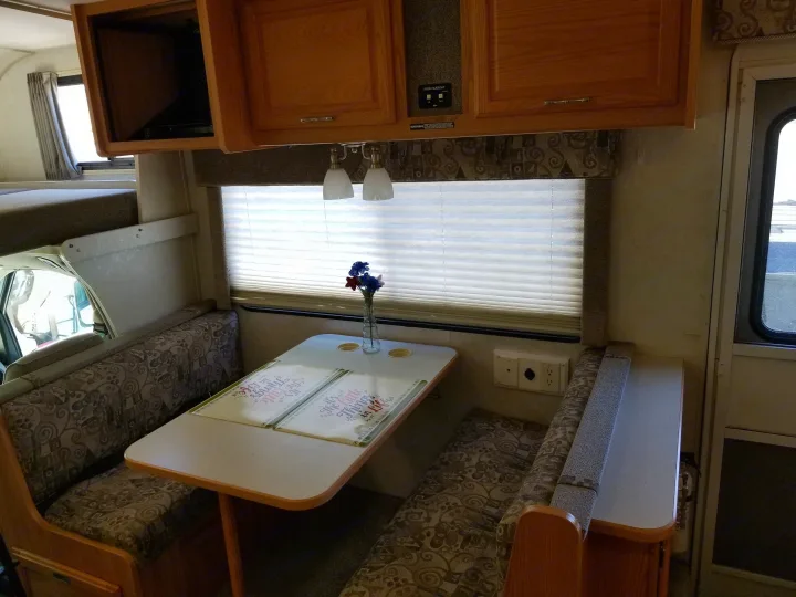 Cozy RV rental interior featuring a dinette with an overhead bunk, wooden cabinets, and a window view, ready for travel.. Winnebago Outlook 2006