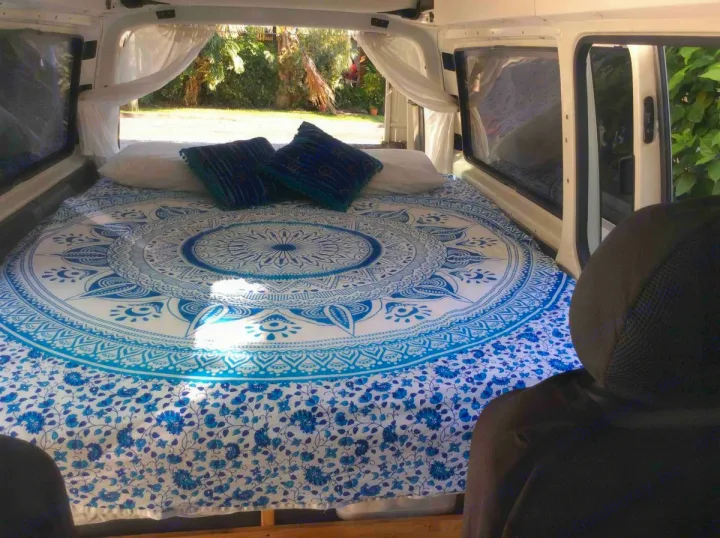 Step into this cozy RV rental! A bed with a blue mandala cover, pillows, and bright outdoor views.. Volkswagen Transporter T5 2005