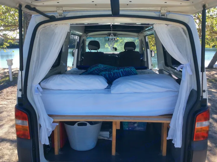 Cozy RV rental campervan interior features a comfortable bed, sheer curtains, and under-bed storage, with a view of water.. Volkswagen Transporter T5 2005