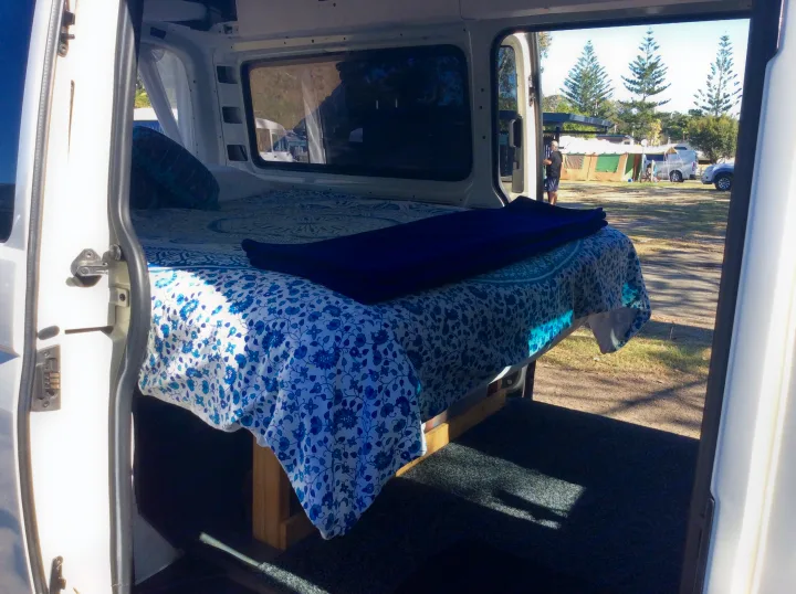 Inside a spacious rv rental campervan, featuring a comfy bed with blue patterns, open to a sunny campsite view.. Volkswagen Transporter T5 2005