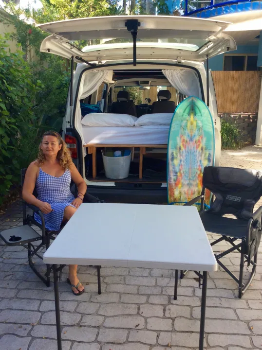 Relaxing by her RV rental: a woman at an outdoor table with her camper van's bed & surfboard inside.. Volkswagen Transporter T5 2005
