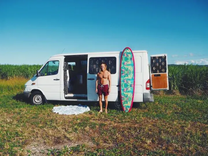 Perfect RV rental for surfers! Man and colorful surfboard stand by a white camper van in a grassy field.. Mercedes-Benz Sprinter 2005