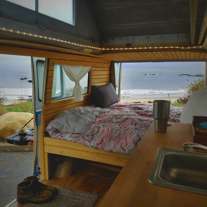 Cozy RV rental with ocean view. Features a bed, small kitchen, outdoor stove, and boots by the door.. Ford Econoline 1966