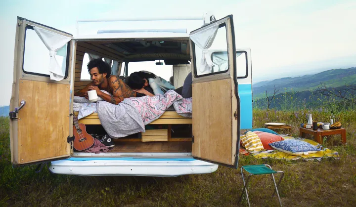 A couple relaxes in their RV rental camper van, doors open to mountain views. A picnic blanket, food, and ukulele are visible.. Ford Econoline 1966