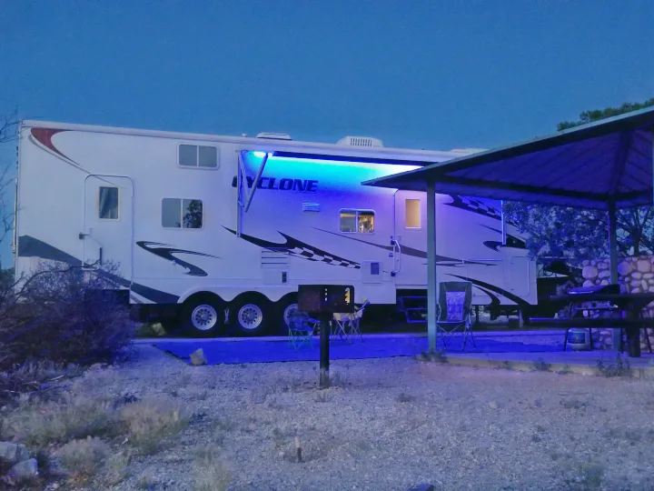 Night scene at an RV rental campsite. A large white travel trailer with glowing blue lights is parked next to a covered picnic area.. Heartland Cyclone 2009