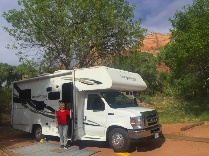 Woman stands by her white Leprechaun rv rental at a scenic campground with green trees and red rock mountains.. Forest River Coachmen Leprechaun 2014