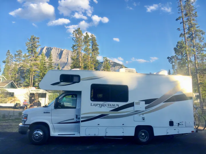 A white Leprechaun Class C RV rental is parked with majestic mountains and pine trees under a clear blue sky.. Forest River Coachmen Leprechaun 2014