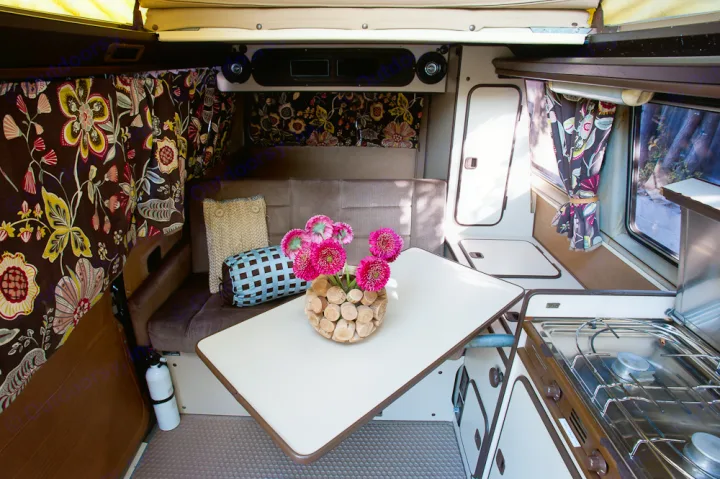 Step inside this cozy RV rental's living space: floral curtains, comfortable seating, a table with pink flowers, and a kitchenette.. Volkswagen Westfalia 1986