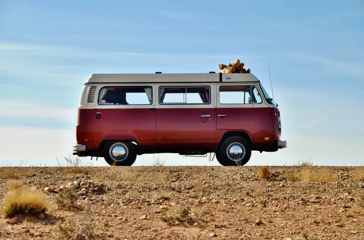 A classic red and white VW camper van, like an RV rental, with logs on its roof, parked on a rocky desert landscape under a blue sky.. Volkswagen Westfalia 1978