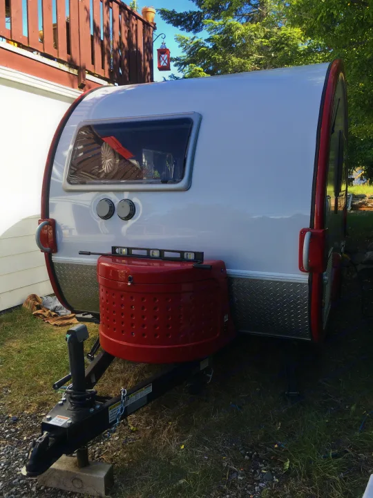 Teardrop RV rental parked in a yard, featuring its front with a red propane tank cover and leveling tool.. T@B Q Floorplan Maxx Package 2016