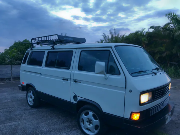 White VW Vanagon with roof rack under a cloudy sky. Perfect for an RV rental getaway!. VW Wolfsburg 1989