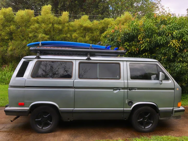 Grey VW T3 camper van with surfboards on roof rack, an ideal RV rental for adventures amidst green landscapes.. VW Wolfsburg 1988