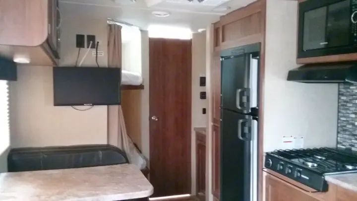 Inside this RV rental: kitchen with stove, microwave, fridge; dining area, TV, and a bunk bed in the background.. Gulfstream 248BH 2018