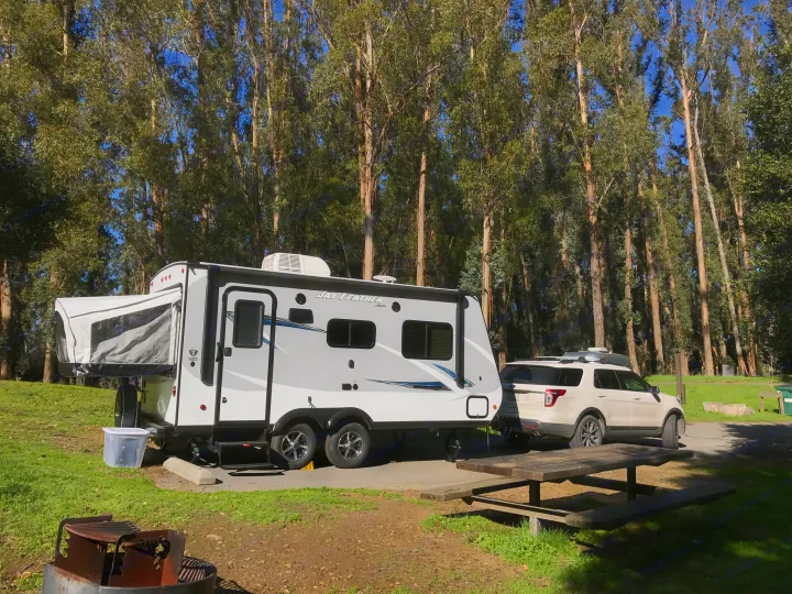 RV rental set up at a sunny campsite with a tow vehicle, picnic table, and tall trees in the background.. Jayco Jay Feather 2017