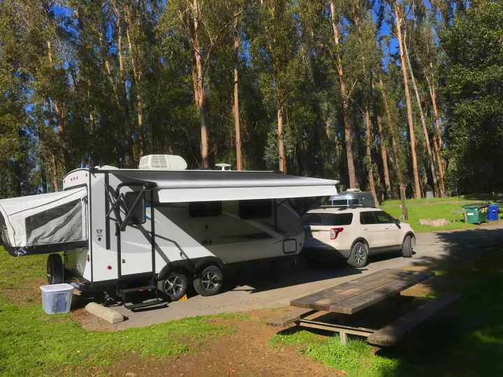 An RV rental travel trailer and its tow SUV are parked in a sunny campground spot with tall trees and a picnic table.. Jayco Jay Feather 2017