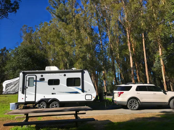 White Jay Feather RV rental travel trailer at a sunny campsite, towed by an SUV, surrounded by tall trees.. Jayco Jay Feather 2017