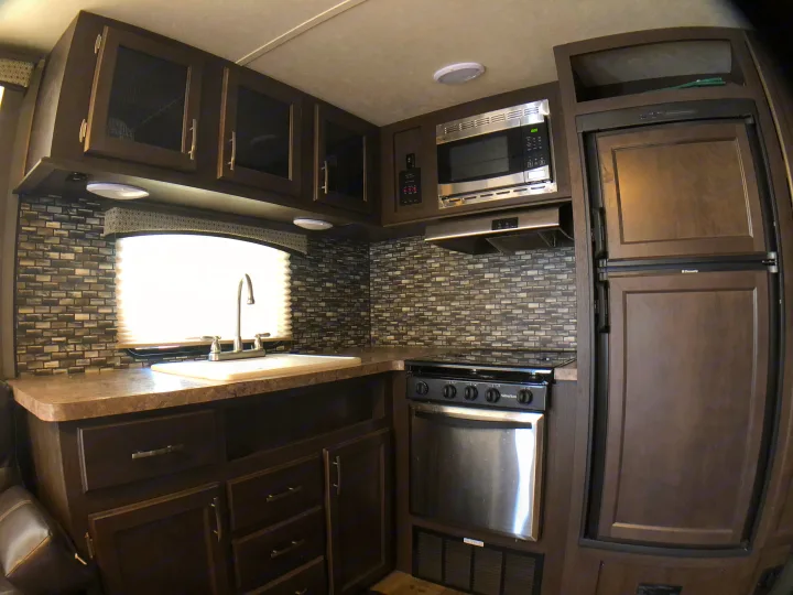 Modern RV rental kitchen with dark wood cabinets, sink, microwave, stove, and refrigerator. Features a tiled backsplash.. Forest River Evo 2017