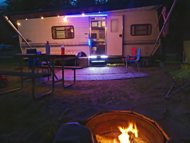 Night at an RV rental campsite. A glowing campfire is in the foreground, with a brightly lit RV and picnic area visible.. Skyline Nomad 1996