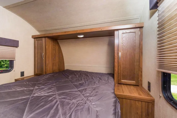 Cozy RV rental bedroom features a bed flanked by wooden storage, overhead cabinet, and windows with blinds.. kz connect 302 2021