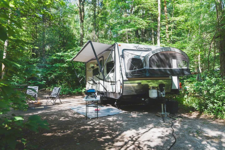 Peaceful rv rental campsite with a travel trailer, chairs, grill, and fire pit in a sunny forest.. Coachmen Freedom Express 2018