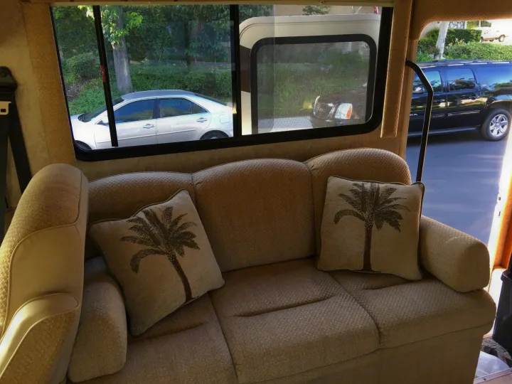 Cozy RV rental interior with a couch, palm tree pillows, and outside view of cars & trees.. National Surf Side 2006