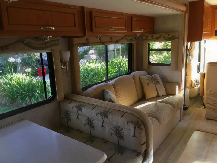 Cozy RV rental interior with a sofa featuring palm tree decor, windows overlooking lush greenery, and wooden cabinets.. National Surf Side 2006