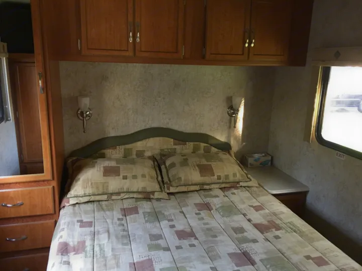 Cozy RV rental bedroom featuring a patterned bed, overhead cabinets, wall sconces, and a side wardrobe for storage.. National Surf Side 2006