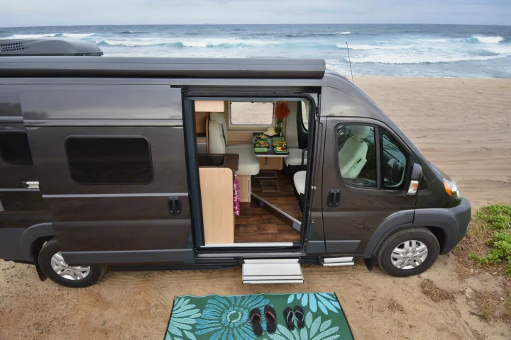 RV rental parked on a sandy beach by the ocean. Its open side door reveals a dining area with food; flip-flops rest on a mat.. Hymer Aktiv 2018