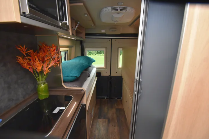 View inside an RV rental: a compact kitchen with a stovetop and flowers, a bed with a blue pillow, and interior windows.. Hymer Aktiv 2018