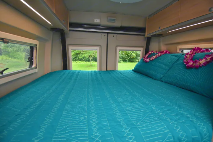 Comfortable RV rental bed with teal bedding, lei, and a serene outdoor view from the windows. Perfect for your travel adventure.. Hymer Aktiv 2018