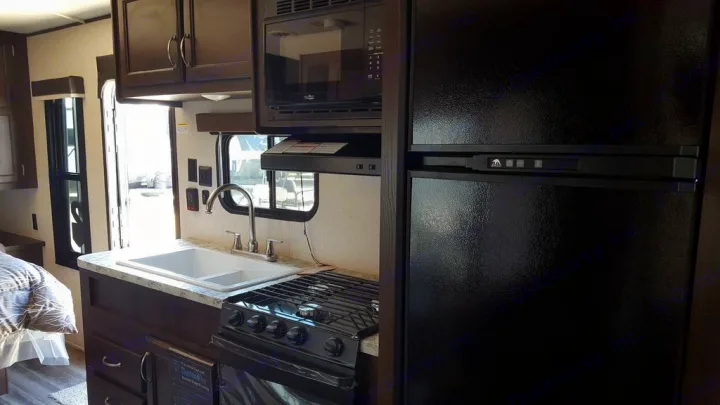 Bright RV rental interior. Modern kitchen with black refrigerator, microwave, stove, and sink. Bedroom area visible.. Jayco Jay Flight 2018