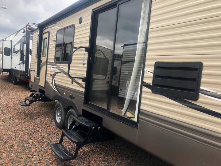 Side view of a modern RV rental, featuring its beige striped exterior, extended black steps, and reflective glass door.. Keystone Hideout 2018