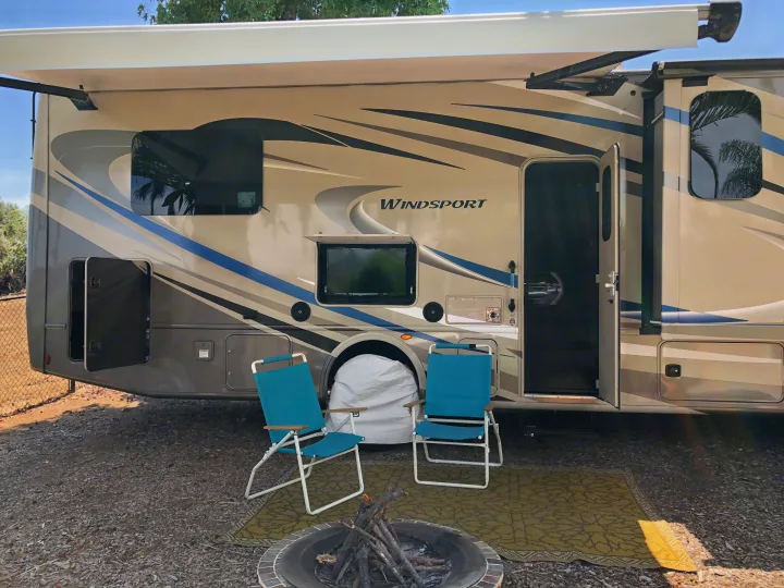 A Windsport RV rental at a sunny campsite, complete with an outdoor TV, chairs, and a fire pit for a relaxing stay.. Thor Windsport 2018