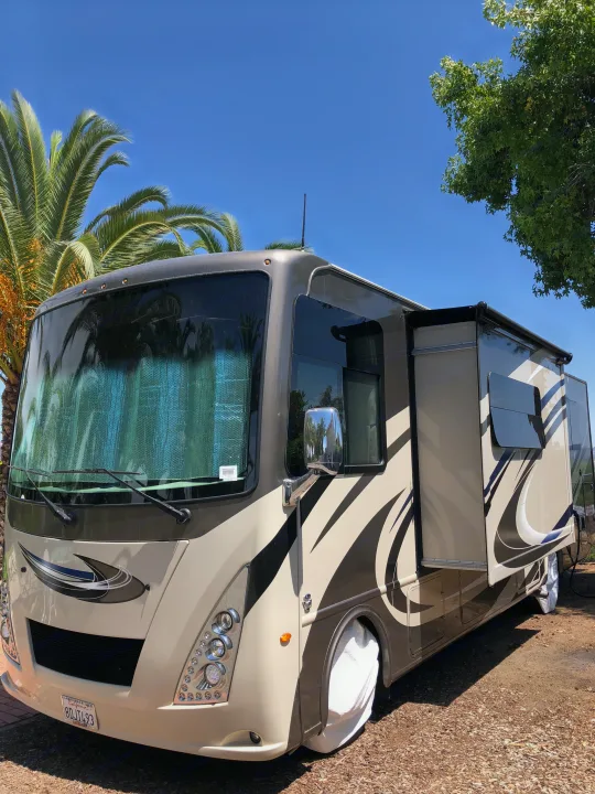 This beige and brown RV rental is parked at a sunny California campsite, featuring an extended slide-out and tire covers.. Thor Windsport 2018