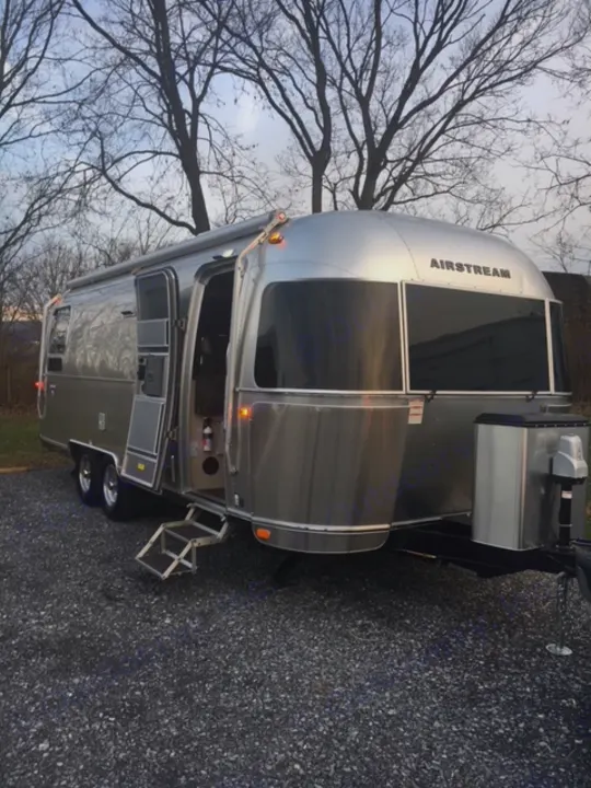 A shiny silver Airstream RV rental, door open & steps extended, parked on gravel with bare trees in background.. Airstream Flying Cloud 2017