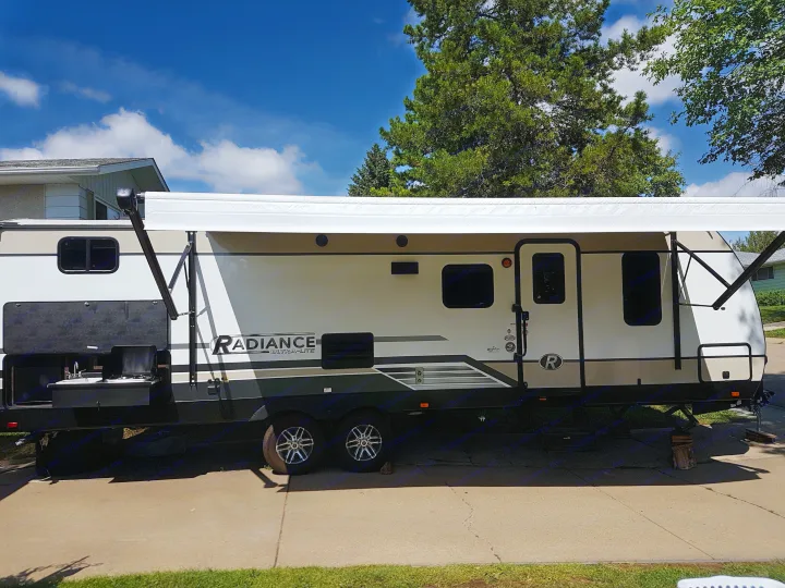 Modern travel trailer RV rental with awning extended, parked on pavement. Blue sky & trees visible above.. Cruiser Rv Corp Radiance 2019
